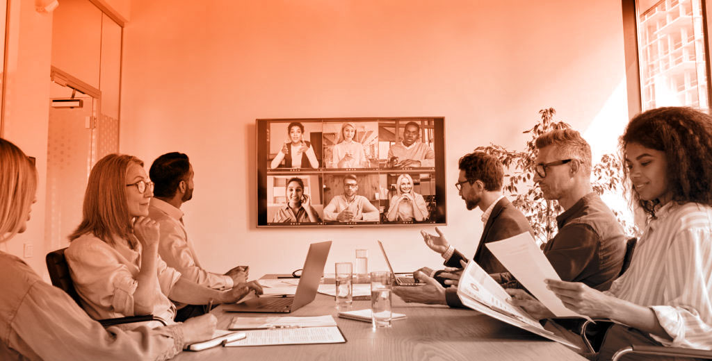 A group of business people sit at a table with a screen showing an online meeting in the background